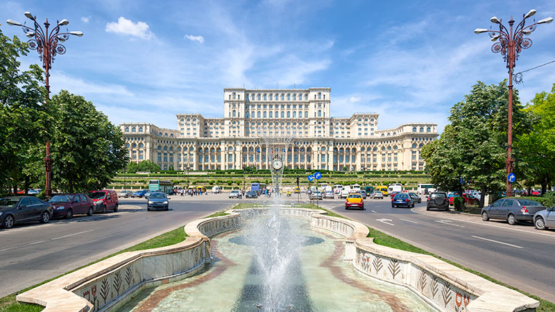 Et enorm palass i Bucuresti med en stor fontene i front. Flere biler p veien rundt fontenen.